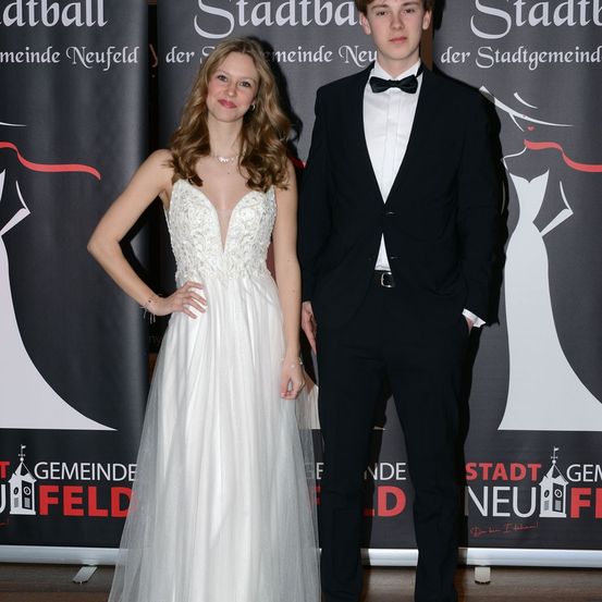 Ein Mann und eine Frau stehen gemeinsam in formeller Kleidung, wahrscheinlich bei einer formellen Veranstaltung. Die Frau trägt ein weißes Kleid und der Mann einen schwarzen Anzug mit Fliege. Sie stehen vor einer Kulisse mit rotem Text.