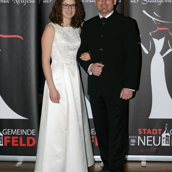 Ein Mann und eine Frau stehen lächelnd zusammen. Die Frau trägt ein weißes Kleid und der Mann einen schwarzen Anzug. Sie stehen vor einer Kulisse mit Text, der 'Gemeinde Feld' und 'Stadt Neufeld' lautet.