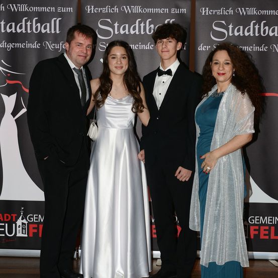 Vier Personen, zwei Männer und zwei Frauen, stehen zusammen. Sie sind alle formell gekleidet, wahrscheinlich bei einer formellen Veranstaltung. Die beiden Frauen tragen Kleider, eines in Silber und das andere in Blau. Die Männer sind in Anzügen. Sie stehen vor einem Banner mit einem Stadtlogo.