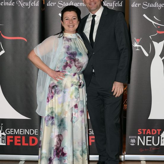 Ein Mann und eine Frau stehen lächelnd zusammen bei einer formellen Veranstaltung. Die Frau trägt ein farbenfrohes Kleid und der Mann einen schwarzen Anzug. Sie stehen vor Bannern mit den Texten 'Neufeld' und 'Feld'.