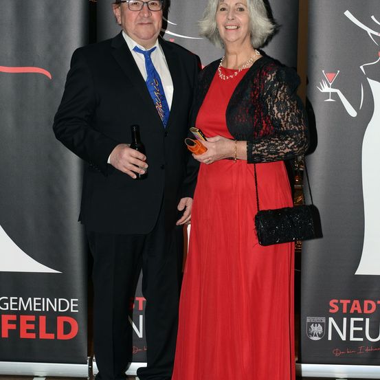 Ein Mann und eine Frau stehen zusammen und lächeln für ein Foto. Der Mann hält eine Flasche und die Frau ein kleines oranges Objekt. Sie tragen formelle Kleidung und stehen vor einem Banner mit den Worten 'Gemeinde Feld'.