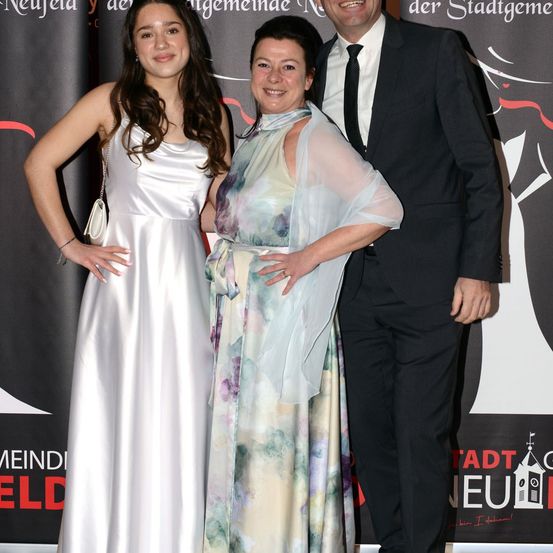 Drei Menschen stehen nebeneinander auf einem roten Teppich. Eine Frau mit langen Haaren lächelt und posiert für ein Foto. Die Frau in der Mitte trägt einen Ring und eine Armbanduhr. Der Mann trägt einen Anzug und eine Krawatte. Hinter ihnen steht eine Wand mit Text.