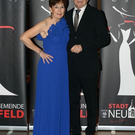 Ein älteres Paar steht vor einem Banner. Die Frau trägt ein blaues Kleid und der Mann einen schwarzen Anzug. Sie scheinen beide für ein Foto zu posieren.