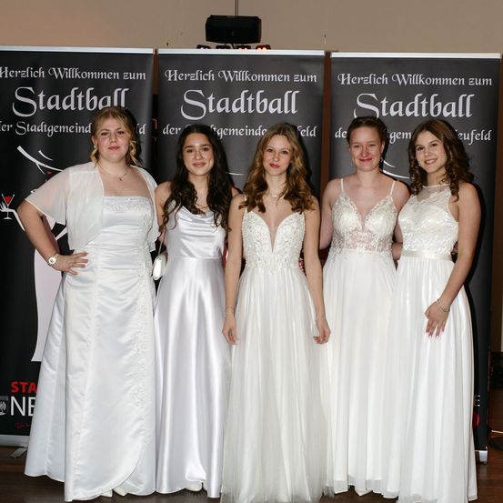 Fünf Frauen in weißen Kleidern stehen nebeneinander vor Bannern, auf denen 'Herzlich Willkommen zum Stadtball' steht. Sie nehmen wahrscheinlich an einer formellen Veranstaltung teil.