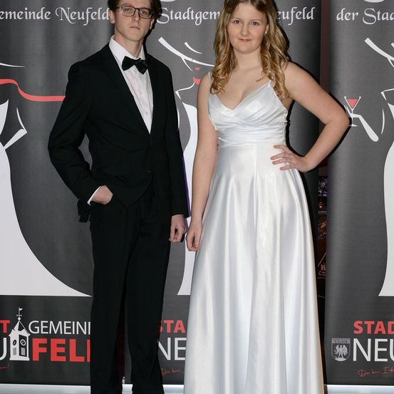 Ein Mann und eine Frau stehen nah beieinander auf einem roten Teppich und lächeln für ein Foto. Sie sind formell gekleidet, und der Hintergrund zeigt einen grauen Banner mit einem stilisierten Design.