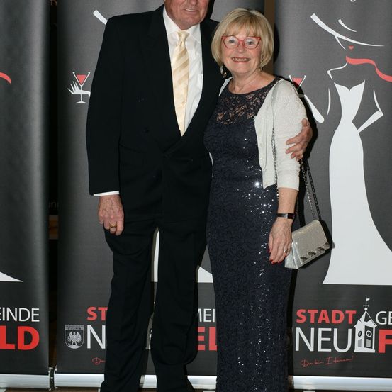 Ein Mann und eine Frau stehen in formeller Kleidung zusammen, lächeln und posieren für ein Foto. Sie stehen vor einem Hintergrund mit einem roten Logo.