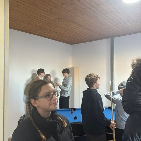 Eine Gruppe von Personen, einige mit Brille, befindet sich in einem Raum mit einem blauen Billardtisch und spielt wahrscheinlich eine Partie Pool.