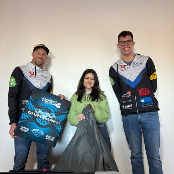 Drei Personen stehen zusammen und halten eine Schachtel und einen Beutel. Der Mann auf der linken Seite trägt einen Hut und hält eine Schachtel. Die Frau in der Mitte hält einen Beutel und lächelt. Der Mann auf der rechten Seite trägt eine Brille und eine Jacke mit Logos.