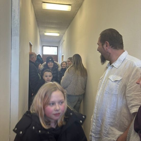 Eine Gruppe von Menschen, darunter eine Frau in einem Pelzmantel und ein Mädchen mit blonden Haaren, geht einen Flur hinunter. Ein Mann mit Bart steht rechts.