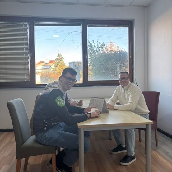 Zwei Männer sitzen an einem Tisch in einem Raum mit Holzboden und Fenstern. Sie scheinen in ein Gespräch vertieft zu sein, während einer einen Laptop betrachtet.