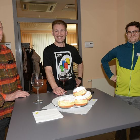 Drei Männer stehen hinter einem Tisch mit Donuts. Einer hält ein Glas Wein. Ein anderer trägt ein schwarzes T-Shirt mit Tetris-Logo.