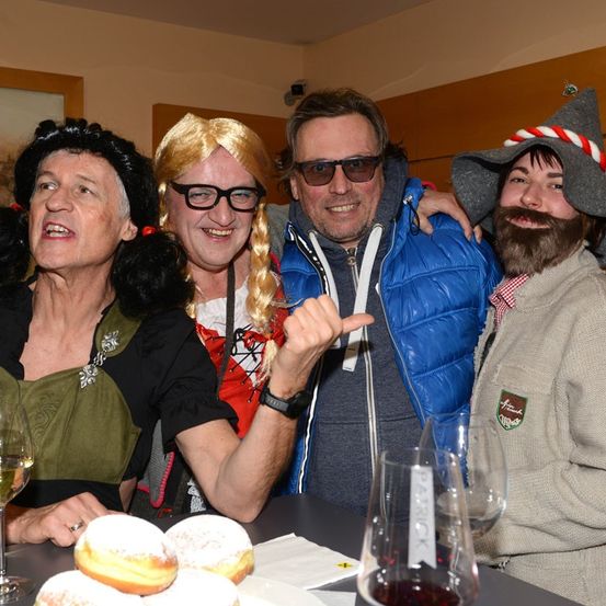 Eine Gruppe von Menschen in Kostümen, einer mit Bart und Hut, sitzen um einen Tisch mit Weingläsern und Donuts.