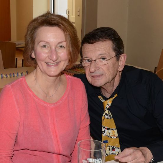 Ein Mann und eine Frau sitzen nah beieinander an einem Tisch. Die Frau trägt ein rosa Hemd und eine Halskette. Der Mann trägt eine Krawatte mit einem Cartoon-Charakter.