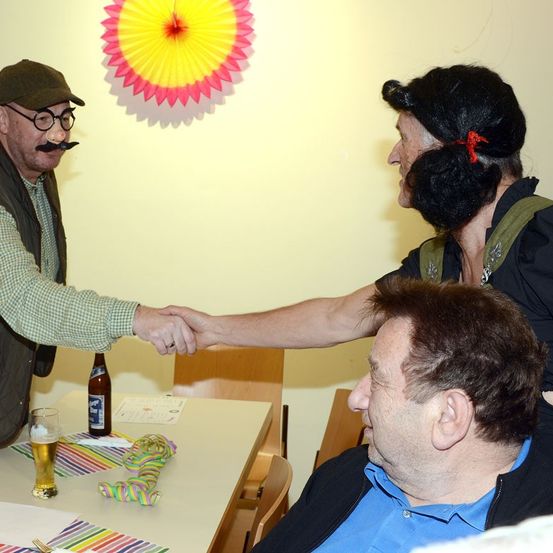 Zwei Männer schütteln sich in einem Raum die Hände. Ein Mann trägt eine grüne Weste, eine Brille und einen Schnurrbart. Der andere Mann trägt eine schwarze Perücke. Hinter ihnen steht ein Tisch mit einem Bier und einem bunten Papierfächer an der Wand.