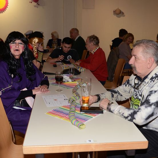 Eine Gruppe von Menschen sitzt an einem langen Tisch in einem Restaurant. Eine Frau in einem lila Umhang schaut einen Mann mit einem Glas Bier an.