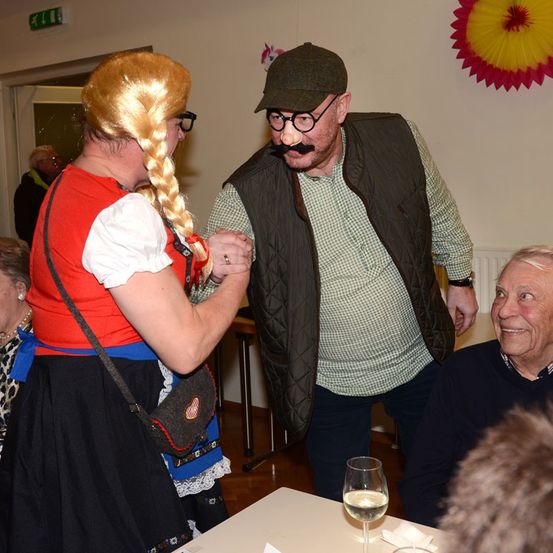 Eine Person in traditioneller bayerischer Kleidung hält die Hände mit einem Mann in Hut und Brille, während ein älterer Mann in der Nähe an einem Tisch mit einem Weinglas sitzt.
