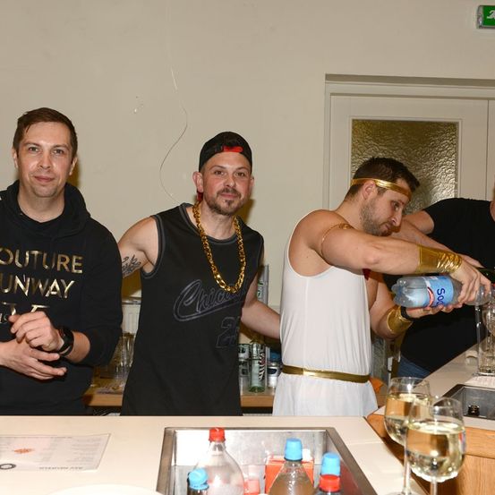 Drei Männer in einer Bar. Einer in einer schwarzen Kapuzenjacke, ein anderer in einem Chicago-Shirt und ein dritter in einer weißen Toga mit einem goldenen Gürtel. Dahinter ein Waschbecken, Flaschen und Weingläser.