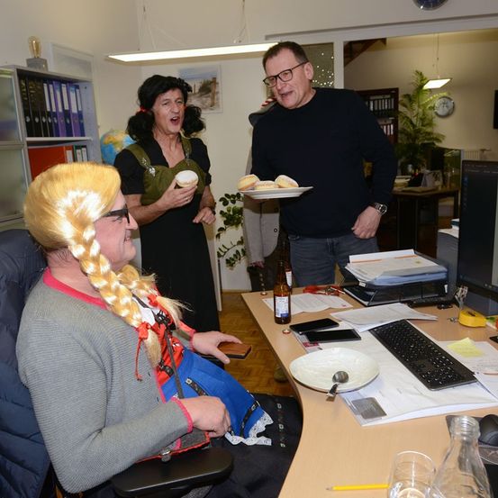 Zwei Erwachsene in Kostümen sind in einem Büro. Einer hält einen Teller mit Gebäck, der andere ein Getränk. Eine Frau mit blonder Perücke schaut sie an. Ein Schreibtisch hat eine Tastatur, einen Teller und eine Flasche.