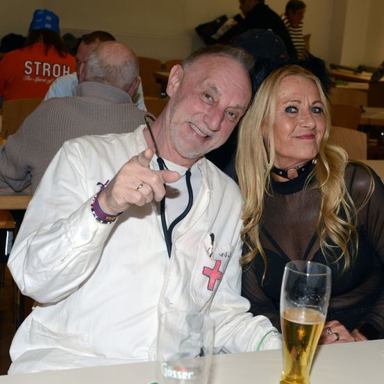 Ein Mann und eine Frau sitzen bei Getränken an einem Tisch, der Mann in einem weißen Mantel und die Frau in einem schwarzen Netzkleid. Sie lächeln beide.