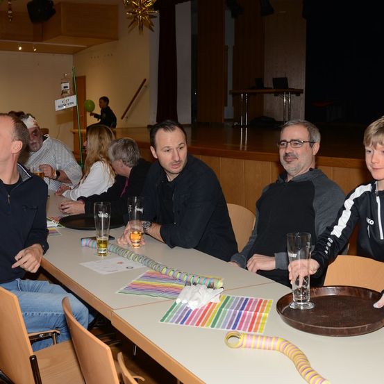 Eine Gruppe von Erwachsenen und einem Kind sitzen mit Gläsern Bier an einem Tisch in einem Saal. Ein Schild mit dem Wort 'EXIT' befindet sich an der Wand. Ein Junge hält ein Glas.