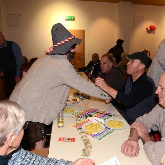 Mehrere ältere Menschen sitzen um einen Tisch, einige tragen Hüte. Ein Mann in einem grauen Hut reicht einem anderen Mann Essen.