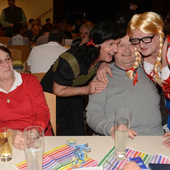 Eine Gruppe von Menschen sitzt an einem Tisch, lächelt und lacht. Ein Mann trägt eine Perücke und eine Brille, eine andere hat ein rotes Band in den Haaren. Sie halten Gläser mit Getränken.