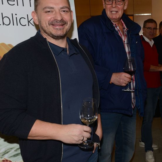 Drei Männer stehen lächelnd eng beieinander, einer hält ein Glas Wein. Ein Banner mit Text ist im Hintergrund zu sehen.