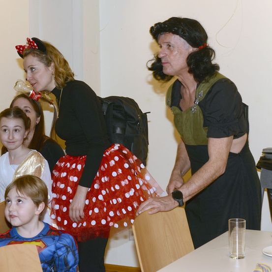 Eine erwachsene Frau in einer Minnie-Maus-Verkleidung berührt den Arm eines Jungen in einer Superman-Verkleidung. Andere Kinder und Erwachsene sind im Hintergrund. Ein Tisch mit Glas und Teller im Vordergrund.