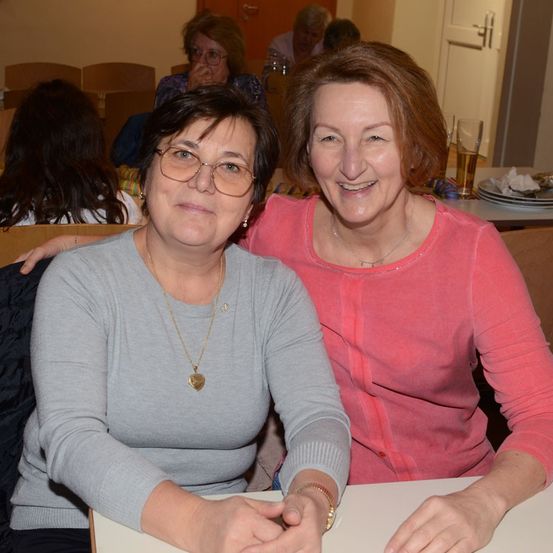 Zwei Frauen sitzen an einem Tisch und lächeln, eine mit Brille und Halskette, die andere in einem rosa Shirt. Hinter ihnen sitzen andere an Tischen, mit Tellern und Gläsern sichtbar.