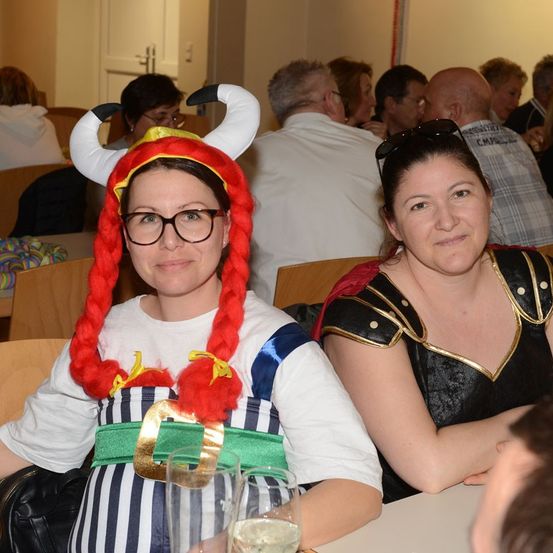 Zwei Frauen sitzen an einem Tisch, eine trägt eine Hörnerhaube und ein Kostüm, die andere ein mittelalterliches Kleid. Sie sitzen nebeneinander, mit anderen Leuten, die hinter ihnen in einem Raum sitzen.