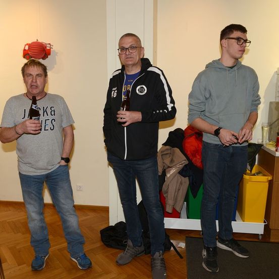 Drei Männer stehen in einem Raum und halten Flaschen. Der Mann auf der linken Seite trägt ein graues T-Shirt, der in der Mitte eine schwarze Jacke und der auf der rechten Seite eine graue Kapuzenjacke. Hinter ihnen hängen Kleidungsstücke an einer Stange.