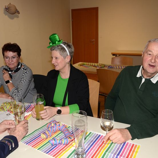 Drei ältere Personen sitzen an einem Tisch und unterhalten sich. Eine Frau in einer grünen Hut hält ein Glas. Hinter ihnen ist eine Tür und eine dekorative Papiergirlande zu sehen.