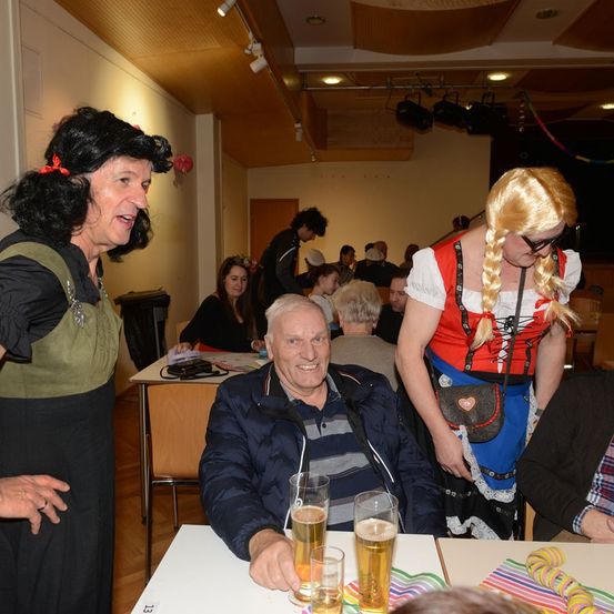Ein Mann und eine Frau in Kostümen stehen neben einem älteren Mann, der Bier trinkt, auf einer Party. Die Party ist drinnen, mit Menschen an Tischen.