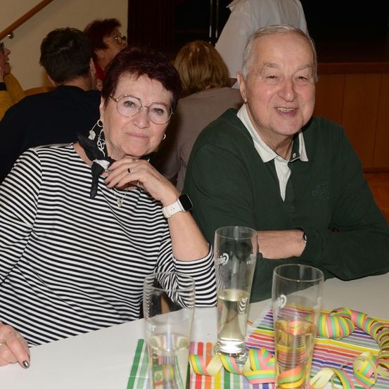 Ein älteres Paar sitzt in einem Restaurant an einem Tisch und lächelt. Sie haben Gläser mit Wasser vor sich, und der Tisch ist mit bunten Luftschlangen dekoriert. Dahinter sitzen andere Leute, und einer steht in der Nähe der Wand.