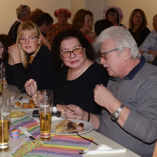 Eine Gruppe von Menschen sitzt um einen Tisch herum, isst und trinkt. Die Frau auf der linken Seite trägt eine Brille und einen Ring. Vor ihr steht ein Glas mit Bier. Der Mann auf der rechten Seite hält eine Gabel und ein Messer. Auf dem Tisch stehen Teller mit Essen, eine Flasche und ein Glas.