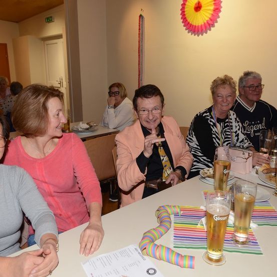 Eine Gruppe von Menschen sitzt an einem Tisch in einem Raum, lächelt und lacht. Ein Mann trägt eine Krawatte mit einer Comicfigur, und der Tisch ist mit Tellern, Gläsern und einem farbenfrohen Strohhalm gedeckt. Eine Wanddekoration verleiht dem Raum eine festliche Note.