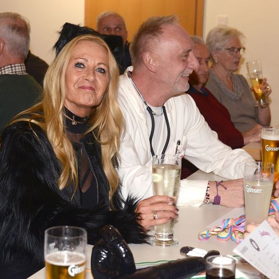 Ein älteres Paar genießt Getränke bei einer Zusammenkunft. Die Frau trägt einen schwarzen Pelzmantel und Katzenohren. Der Mann hat eine Stethoskop um den Hals. Hinter ihnen sitzen und plaudern Menschen, einige halten Getränke.