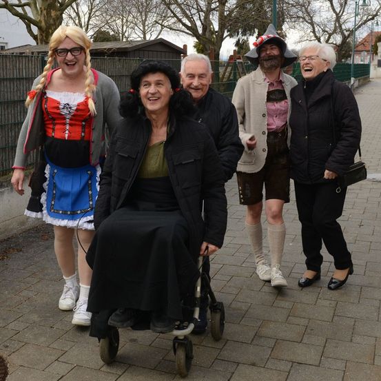 Fünf Personen, darunter eine Frau im Rollstuhl, posieren in traditioneller Kleidung für ein Foto. Bäume und ein Zaun sind im Hintergrund zu sehen.