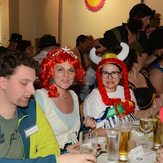 Mehrere Menschen in einem Restaurant sind in Kostümen gekleidet. Einer trägt einen hornbewehrten Wikingerhelm. Eine andere trägt eine rote Perücke und eine Perlenkette. Ein Mann neben ihr trägt ein Namensschild. Gläser, Wein und Biergläser stehen auf dem Tisch.