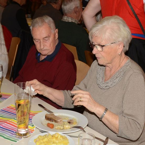 Ein älteres Paar isst in einem Restaurant, der Mann mit einem Taschentuch in der Hand, die Frau mit einer Brille und einem Ring. Sie sitzen neben einem Glas Bier und einem Teller mit Fleisch und Gemüse. Andere Gäste sind an benachbarten Tischen platziert.