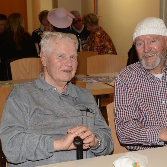 Zwei ältere Männer lächeln in die Kamera und sitzen in einem Essbereich, mit anderen Menschen im Hintergrund. Ein Mann hat einen Stock und ein graues Hemd, während der andere einen weißen Hut und ein kariertes Hemd trägt.