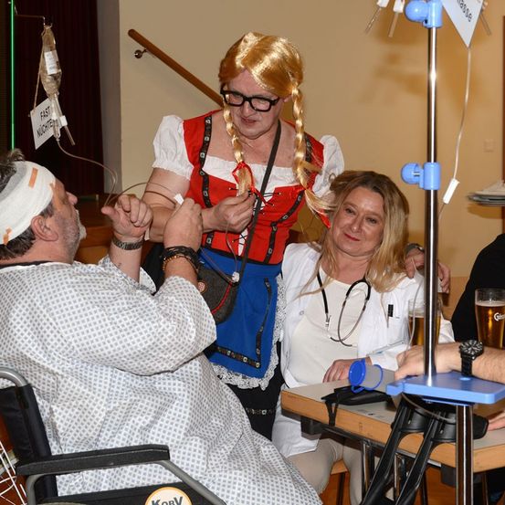 Eine Frau in einem Kostüm steht neben einem Mann im Rollstuhl, während eine Ärztin sie untersucht. Sie befinden sich in einer Krankenhausumgebung, mit einer medizinischen Infusionsflasche und einem Schild, das Fast Nüchten steht. Eine andere Person hält ein Glas Bier.