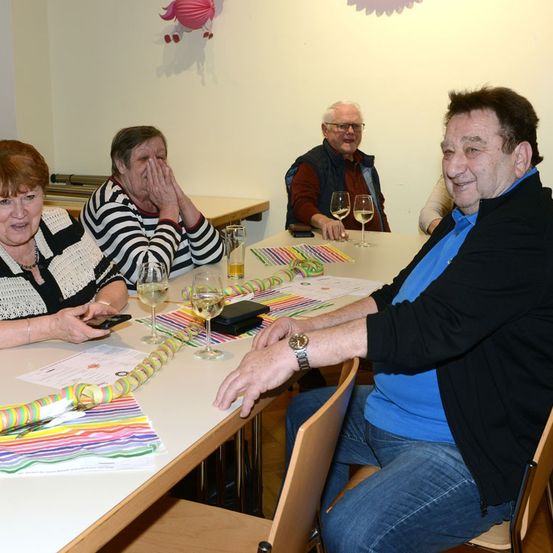 Vier Erwachsene sitzen an einem Tisch mit Weinläsern und bunten Bändern. Ein Mann lächelt, während die anderen überrascht aussehen.
