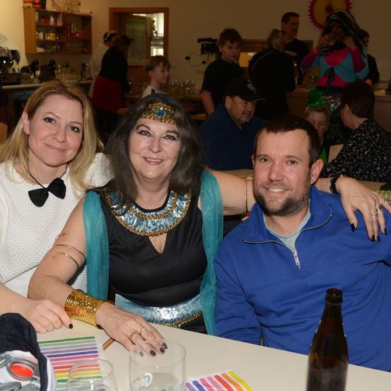 Zwei Frauen und ein Mann sitzen mit bunten Stiften und einer Flasche am Tisch und lächeln. Sie tragen festliche Kostüme. Im Hintergrund unterhalten sich Menschen und eine Frau trinkt aus einem Glas.
