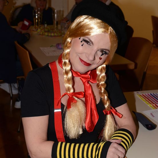 Eine Frau in Clown-Kostüm mit rotem Bogen und gestreiften Ärmeln posiert für ein Foto. Sie lächelt mit verschränkten Armen.