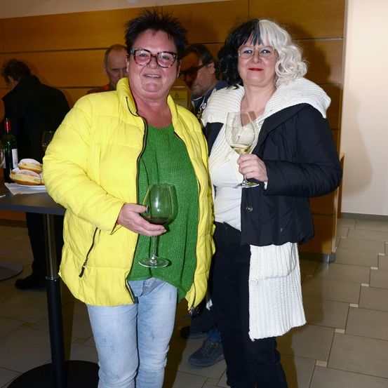 Zwei Frauen stehen drinnen bei einer Veranstaltung, eine in einer gelben Steppjacke, die andere in einer schwarzen Jacke mit weißem Schal, beide halten Weingläser. Ein Tisch mit Gebäck und Weinflaschen steht hinter ihnen, mit anderen Leuten im Hintergrund.