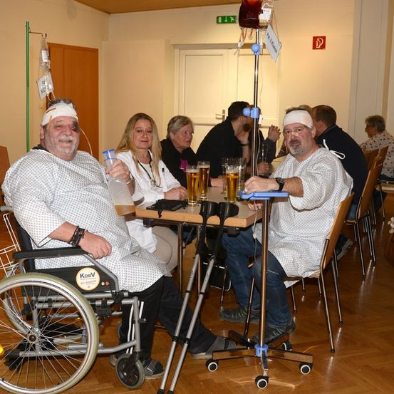 Eine Gruppe von Menschen in Krankenhauskitteln sitzt um einen Tisch mit Getränken. Ein Mann ist im Rollstuhl und ein anderer hält eine Infusionsflasche.