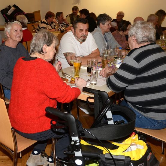 Eine Gruppe von Menschen sitzt an Tischen in einem Raum, einige mit Biergläsern. Eine ältere Frau in einem roten Pullover isst und ein Kinderwagen ist in der Nähe.