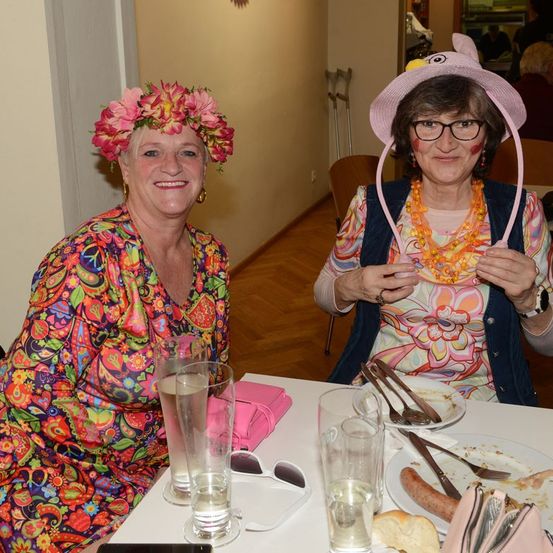 Zwei Frauen in bunten Kleidern und Hüten sitzen an einem Tisch. Eine trägt einen Blumenkranz, die andere einen rosa Hut. Der Tisch ist mit Essen, Gläsern und Besteck gedeckt. Der Hintergrund zeigt eine Wand und Stühle.