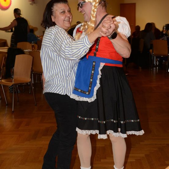 Zwei Frauen tanzen auf einem Holzboden. Eine trägt ein traditionelles Kleid, die andere ein gestreiftes Hemd. Sie sind umgeben von Tischen, Stühlen und einigen Leuten. Die Wände sind weiß mit dekorativen Elementen.